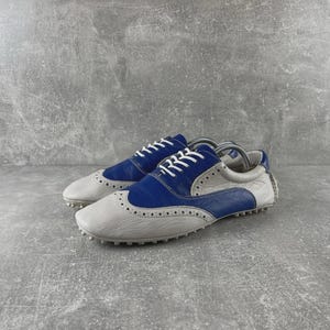 May include: A pair of two-tone golf shoes. The shoes are grey and blue with white laces. The shoes have a textured sole and a classic wingtip design. The shoes are made of leather.