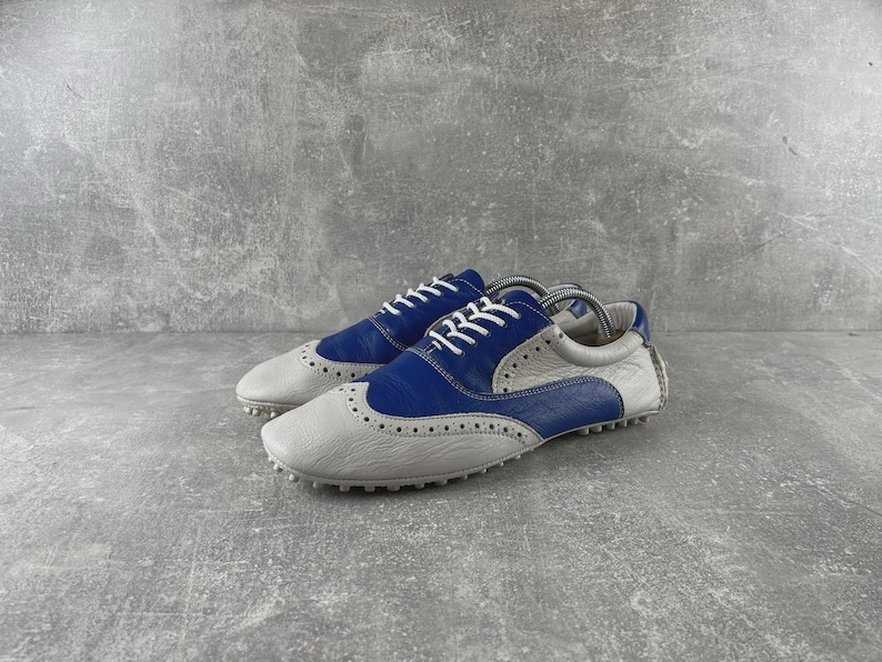 May include: A pair of two-tone golf shoes. The shoes are grey and blue with white laces. The shoes have a textured sole and a classic wingtip design. The shoes are made of leather.