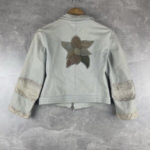 Peut inclure: Veste bleu clair avec un motif floral au dos. La veste a une fermeture &eacute;clair et des patchs d&eacute;coratifs sur les manches. Le motif floral est compos&eacute; de diff&eacute;rentes nuances de marron, de vert et de gris.