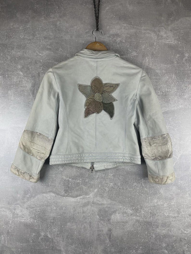 Peut inclure: Veste bleu clair avec un motif floral au dos. La veste a une fermeture &eacute;clair et des patchs d&eacute;coratifs sur les manches. Le motif floral est compos&eacute; de diff&eacute;rentes nuances de marron, de vert et de gris.