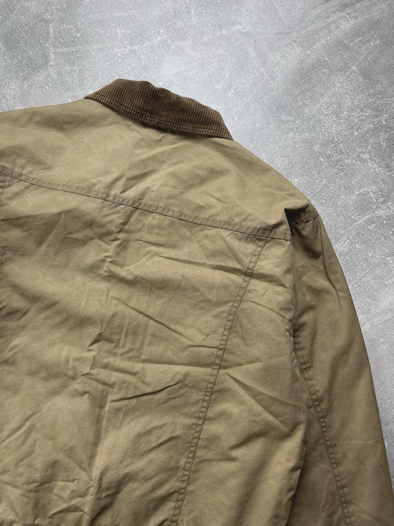 Puede incluir: Una chaqueta de color tostado con cuello de pana marr&oacute;n y una costura horizontal en la parte superior de la espalda. La prenda est&aacute; confeccionada en un tejido liso y mate con detalles de costuras visibles en los hombros y la espalda.