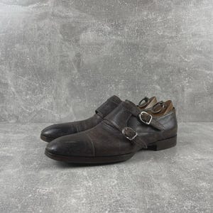 May include: A pair of brown leather double monk strap shoes with silver buckles. The shoes are displayed on a gray surface. The shoes have a classic design and are suitable for formal occasions.