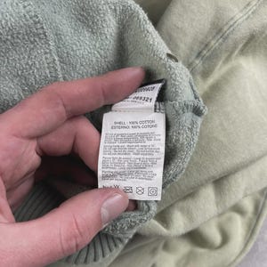 May include: A close-up shot of a light green cotton garment with a care label. The label details fabric composition, stating "SHELL: 100% COTTON" and "ESTERNO: 100% COTONE". The garment is likely a sweatshirt or similar clothing item.
