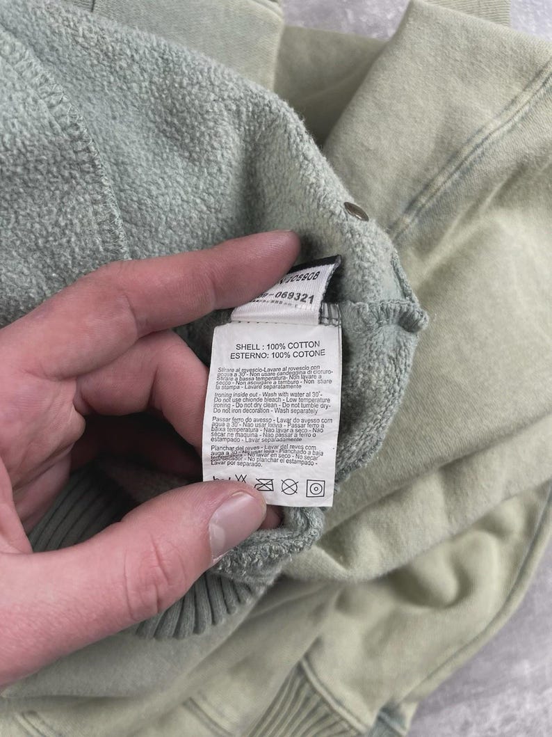 May include: A close-up shot of a light green cotton garment with a care label. The label details fabric composition, stating "SHELL: 100% COTTON" and "ESTERNO: 100% COTONE". The garment is likely a sweatshirt or similar clothing item.