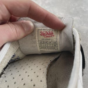 May include: The interior of a white Von Dutch sneaker featuring a size label that reads US 8, EUR 41, UK 7, and 26 cm. The label includes the brand logo, website, and states Made in China. The insole is white with small ventilation holes.