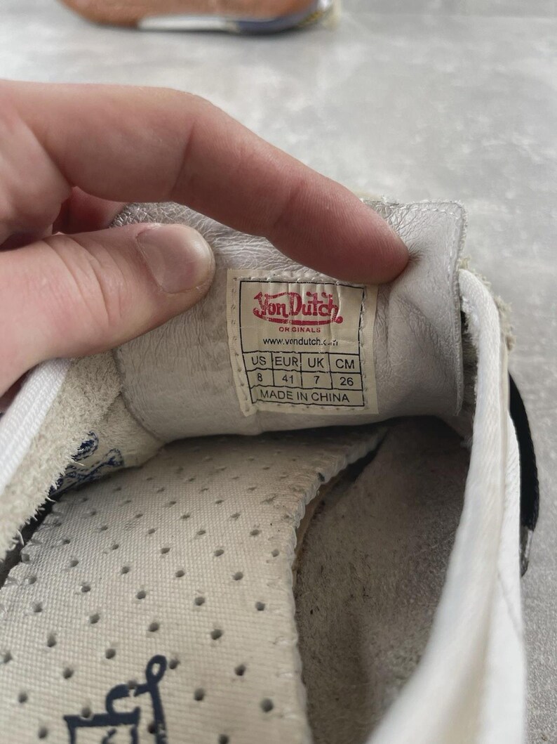 May include: The interior of a white Von Dutch sneaker featuring a size label that reads US 8, EUR 41, UK 7, and 26 cm. The label includes the brand logo, website, and states Made in China. The insole is white with small ventilation holes.
