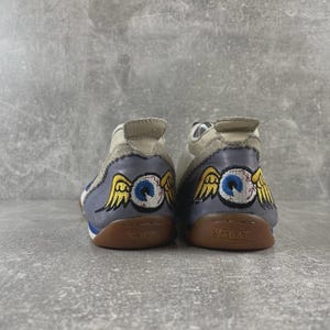 May include: A pair of low-top sneakers featuring a light gray upper and a blue heel panel. The heel displays an embroidered graphic of a blue eyeball with yellow wings. The gum rubber soles are embossed with the Von Dutch brand name on the back.