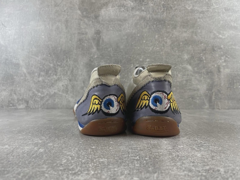 May include: A pair of low-top sneakers featuring a light gray upper and a blue heel panel. The heel displays an embroidered graphic of a blue eyeball with yellow wings. The gum rubber soles are embossed with the Von Dutch brand name on the back.