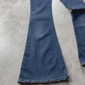 May include: A pair of dark blue denim jeans featuring a flared leg silhouette. The fabric displays a subtle whiskered fading pattern around the upper thigh area, with a classic stitched hem at the bottom of the wide leg opening.