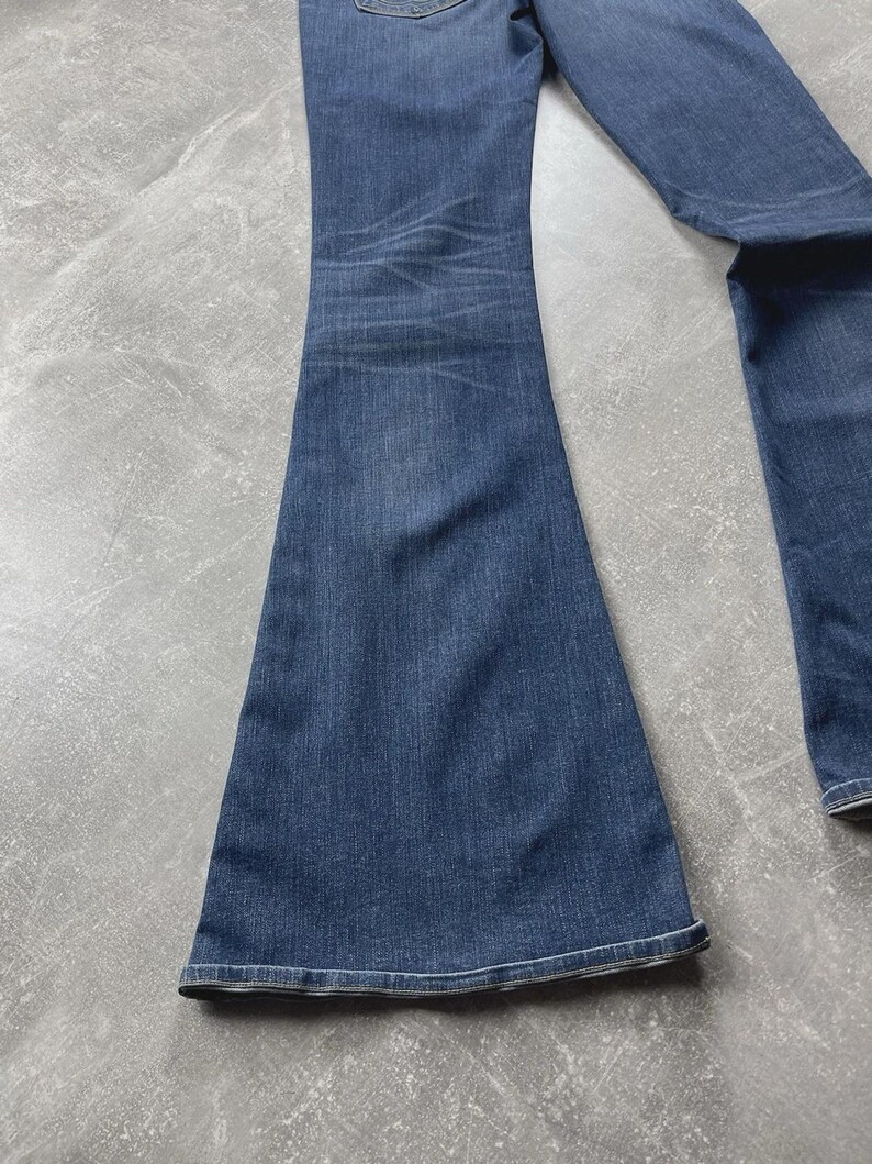 May include: A pair of dark blue denim jeans featuring a flared leg silhouette. The fabric displays a subtle whiskered fading pattern around the upper thigh area, with a classic stitched hem at the bottom of the wide leg opening.