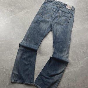 May include: Blue denim jeans with a classic five-pocket design. The jeans feature a faded wash and a flared leg opening. The back pockets have a signature stitch design. A red tag is visible on the back pocket.