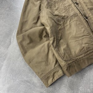 Puede incluir: Una chaqueta utilitaria de color tostado con cierre frontal de botones, cuello en punta y dos grandes bolsillos de parche en el pecho. La prenda cuenta con mangas largas, pu&ntilde;os abotonados y detalles de costuras visibles a lo largo de los paneles.