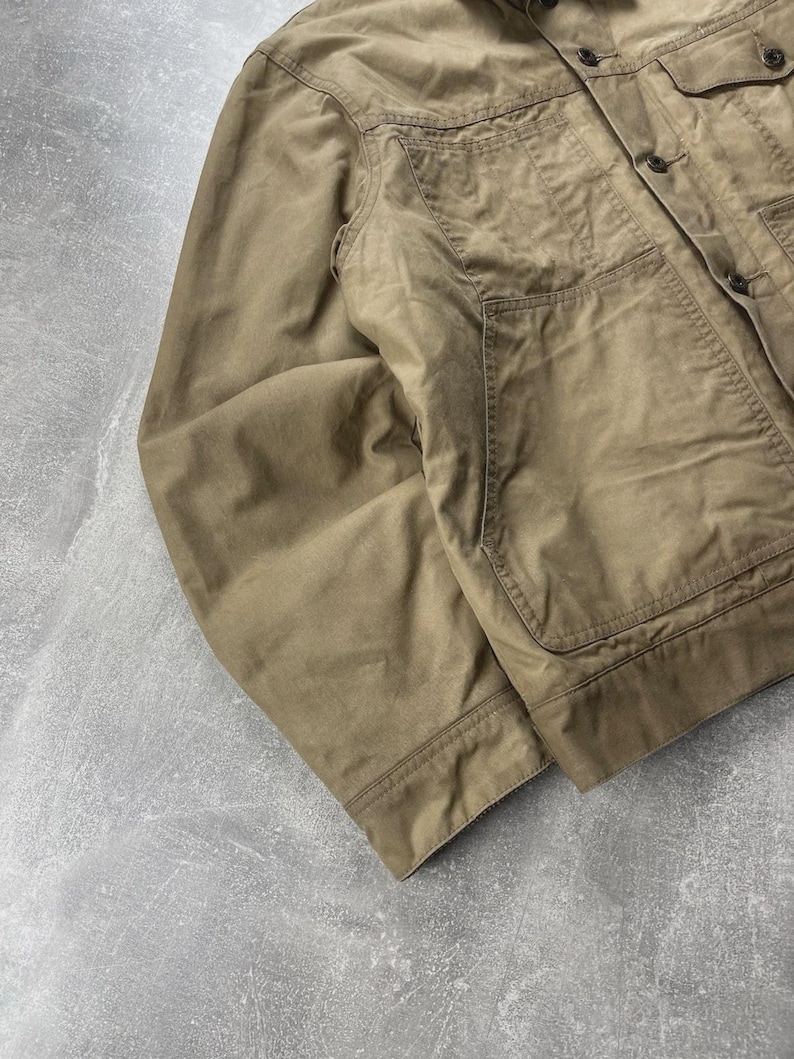 Puede incluir: Una chaqueta utilitaria de color tostado con cierre frontal de botones, cuello en punta y dos grandes bolsillos de parche en el pecho. La prenda cuenta con mangas largas, pu&ntilde;os abotonados y detalles de costuras visibles a lo largo de los paneles.
