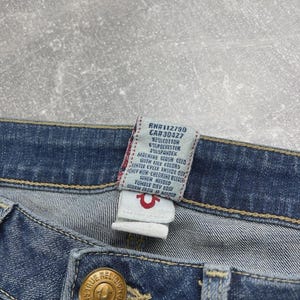 May include: Blue denim jeans featuring a gold-toned metal button embossed with the True Religion brand logo. A white fabric care tag is attached to the interior waistband, displaying text including RN#112790, CA#30427, 92% cotton, 6% polyester, and 2% spandex.