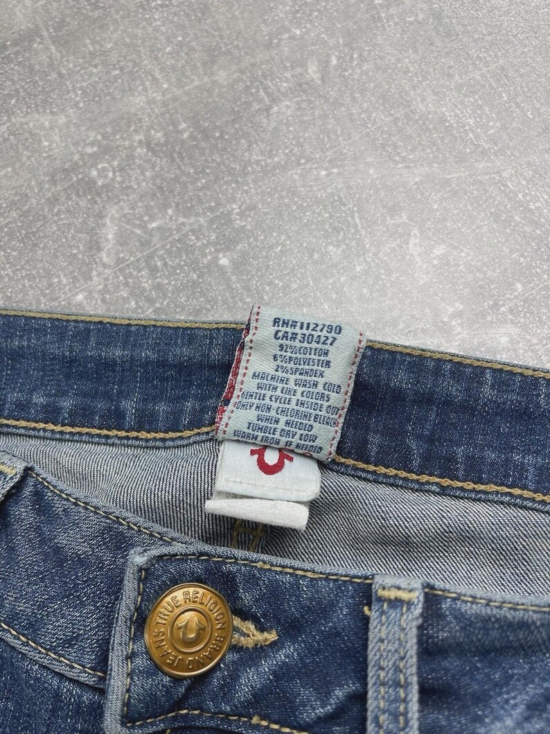 May include: Blue denim jeans featuring a gold-toned metal button embossed with the True Religion brand logo. A white fabric care tag is attached to the interior waistband, displaying text including RN#112790, CA#30427, 92% cotton, 6% polyester, and 2% spandex.