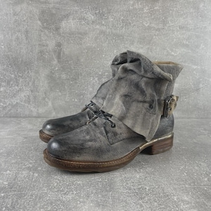 May include: A distressed grey leather ankle boot featuring a slouchy, folded cuff design, a side buckle closure, dark laces, and a low brown stacked heel. The leather has a textured, weathered finish with darker shading around the toe and heel.