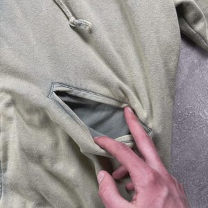 May include: A pale green hoodie featuring a kangaroo pocket and a drawstring hood. The hoodie is made from a soft, textured material and has a faded look. The pocket is open, showing the inside lining.