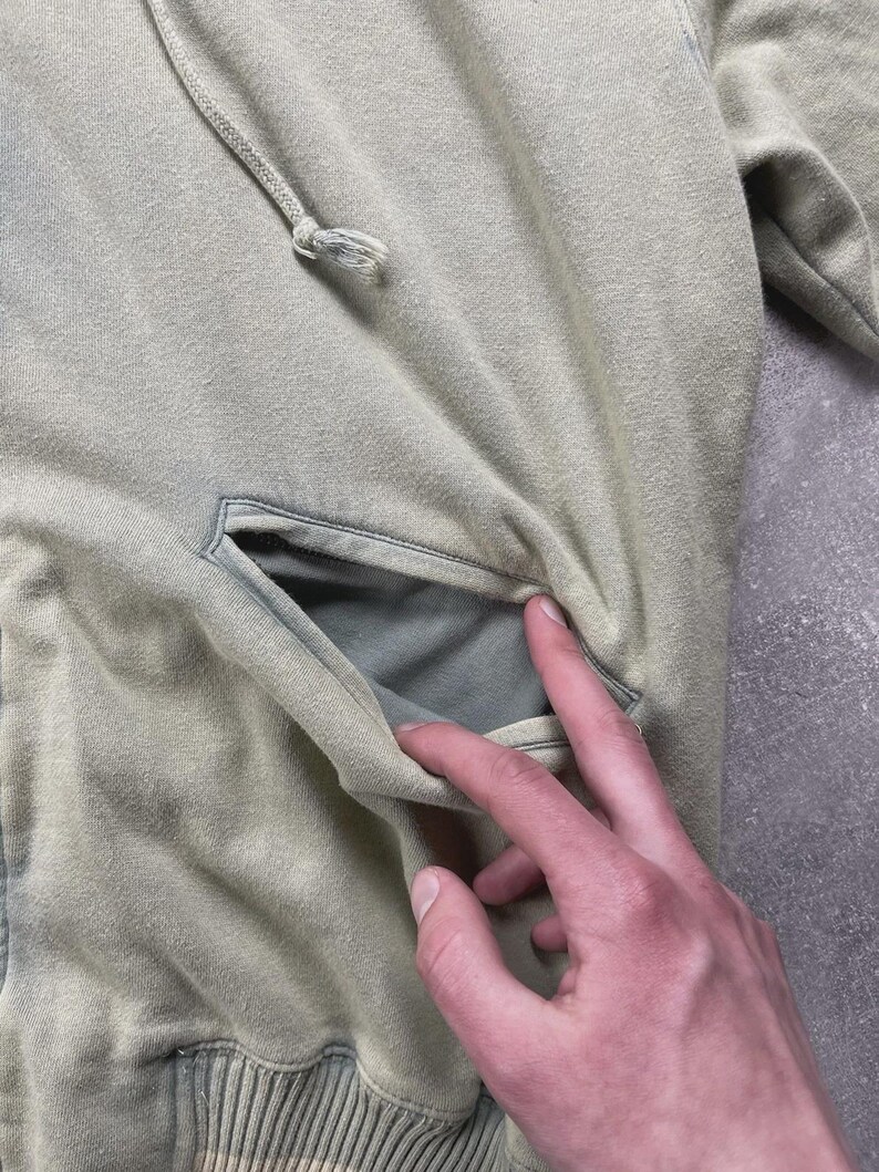 May include: A pale green hoodie featuring a kangaroo pocket and a drawstring hood. The hoodie is made from a soft, textured material and has a faded look. The pocket is open, showing the inside lining.
