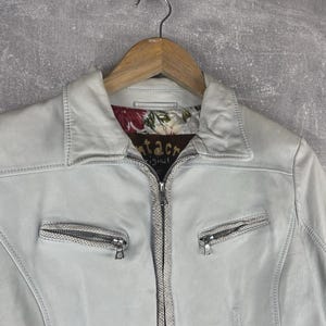 Peut inclure: Veste en cuir bleu clair avec un col et une fermeture &eacute;clair. La veste a deux poches zipp&eacute;es et une doublure &agrave; motifs floraux. Le nom de la marque "tacre original" est visible sur la doublure.