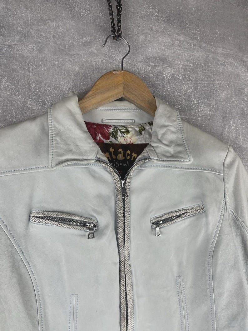 Peut inclure: Veste en cuir bleu clair avec un col et une fermeture &eacute;clair. La veste a deux poches zipp&eacute;es et une doublure &agrave; motifs floraux. Le nom de la marque "tacre original" est visible sur la doublure.