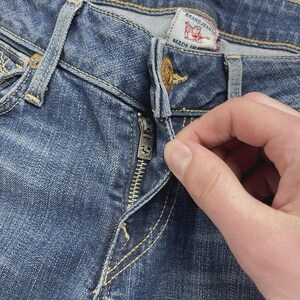 May include: Medium blue denim jeans featuring a metal button closure and a silver-toned zipper fly. The interior waistband displays a white fabric label with red text reading BRAND JEANS and a red horseshoe logo, with MADE IN visible below.