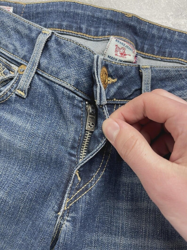 May include: Medium blue denim jeans featuring a metal button closure and a silver-toned zipper fly. The interior waistband displays a white fabric label with red text reading BRAND JEANS and a red horseshoe logo, with MADE IN visible below.