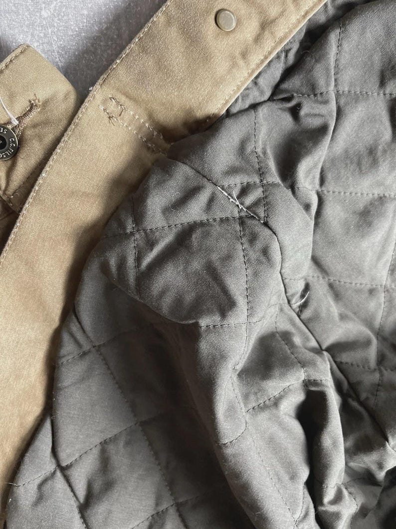 Puede incluir: Una chaqueta de color tostado con un forro acolchado gris oscuro con patr&oacute;n de diamantes. La capa exterior es de tela tostada s&oacute;lida con un cierre de bot&oacute;n a presi&oacute;n met&aacute;lico visible cerca del cuello. Un peque&ntilde;o bot&oacute;n met&aacute;lico est&aacute; a la izquierda.