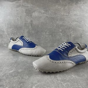 May include: A pair of white and blue leather golf shoes with white laces. The shoes have a wingtip design with small perforations and a rubber sole. The shoes are designed for golf.