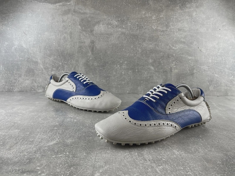 May include: A pair of white and blue leather golf shoes with white laces. The shoes have a wingtip design with small perforations and a rubber sole. The shoes are designed for golf.
