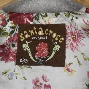 Peut inclure: Gros plan sur un tissu &agrave; motifs floraux avec une &eacute;tiquette marron. L'&eacute;tiquette indique "Santa Croce original garment dyed" et pr&eacute;sente une rose rouge peinte. Le tissu a un fond blanc avec des fleurs roses et rouges.