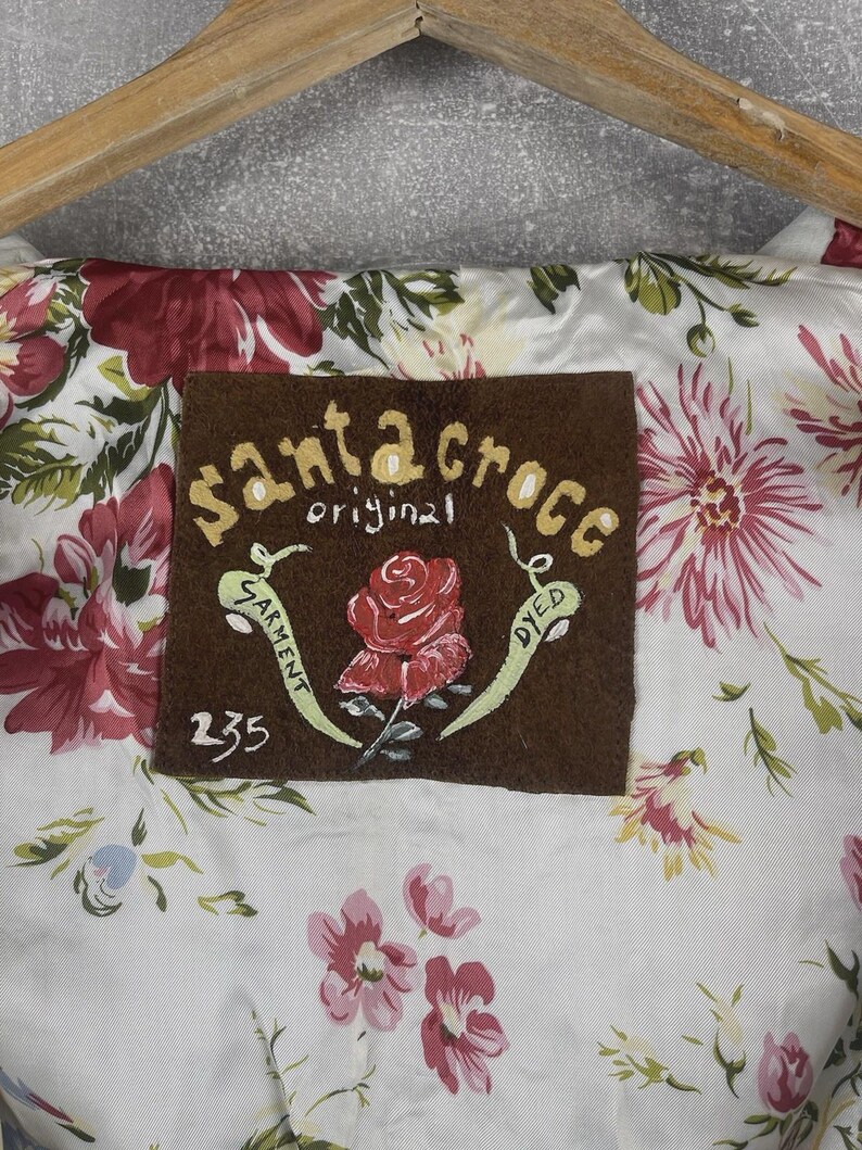 Peut inclure: Gros plan sur un tissu &agrave; motifs floraux avec une &eacute;tiquette marron. L'&eacute;tiquette indique "Santa Croce original garment dyed" et pr&eacute;sente une rose rouge peinte. Le tissu a un fond blanc avec des fleurs roses et rouges.