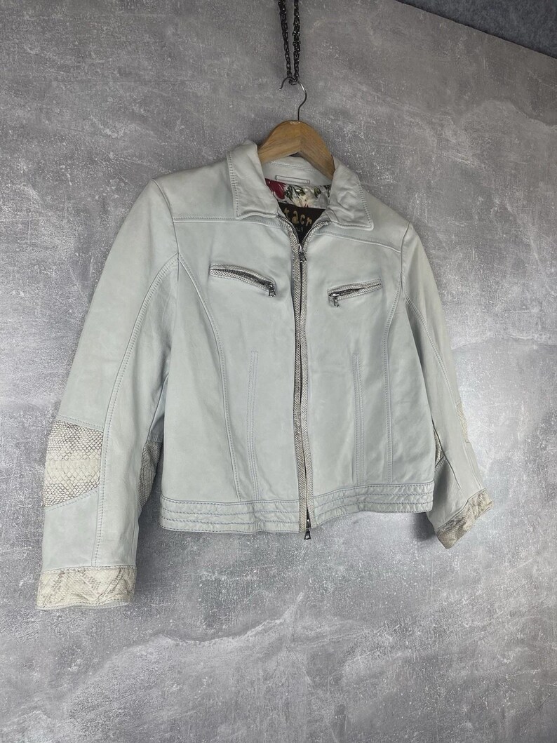 Peut inclure: Veste en cuir gris clair avec fermeture &eacute;clair et deux poches zipp&eacute;es. La veste a un col et des coutures d&eacute;coratives. Les manches pr&eacute;sentent un motif peau de serpent sur les coudes et les poignets. La doublure int&eacute;rieure a un motif floral.