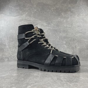 May include: A black suede ankle boot featuring a cage-like design with smooth black leather straps across the toe and sides. The boot includes light gray laces, a high-top silhouette, and a thick, black lugged rubber sole with a defined heel.