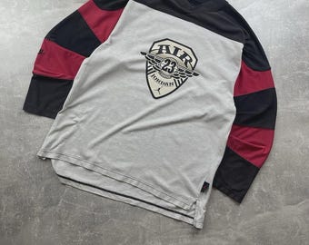 May include: A long-sleeved shirt with a gray body, black sleeves, and red accents. The shirt features a shield-shaped graphic with the text "AIR 23 JORDAN" in black and white. The shirt is made of a soft material.
