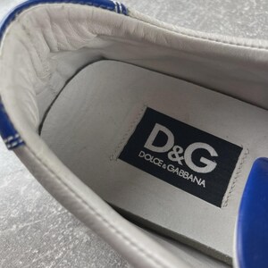 May include: Close-up of a white leather sneaker interior with blue trim. The insole features a black label with the white text "D&G DOLCE & GABBANA". The shoe's interior is white leather.