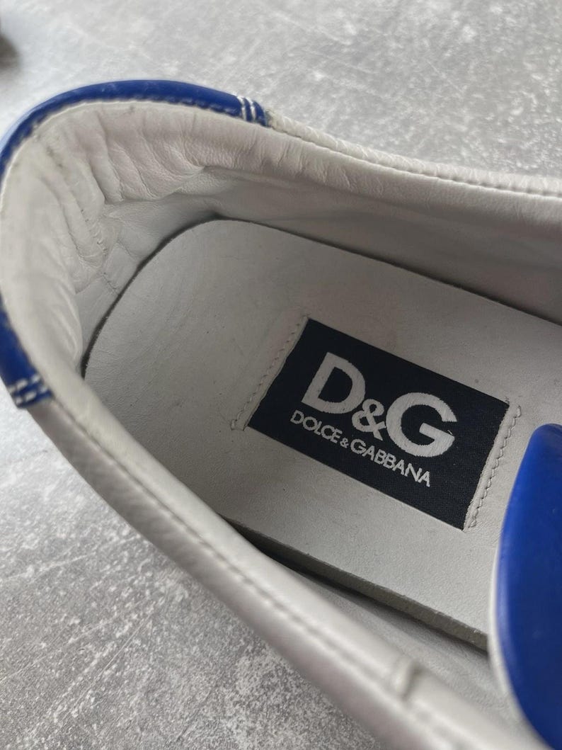 May include: Close-up of a white leather sneaker interior with blue trim. The insole features a black label with the white text "D&G DOLCE & GABBANA". The shoe's interior is white leather.