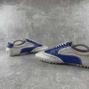 May include: A pair of white and blue leather golf shoes. The shoes have a classic brogue design with white laces and a rubber sole. The heel features a blue accent with a logo. Designed for comfort and performance on the golf course.