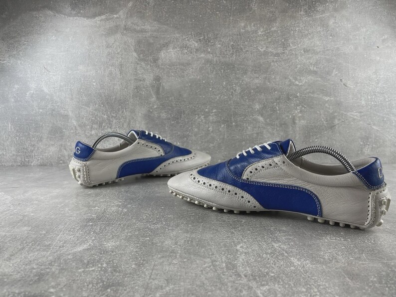 May include: A pair of white and blue leather golf shoes. The shoes have a classic brogue design with white laces and a rubber sole. The heel features a blue accent with a logo. Designed for comfort and performance on the golf course.