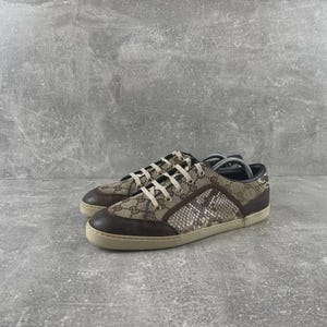 Op de afbeelding: Lage sneakers met een bovenwerk van beige en bruin monogram canvas met donkerbruine leren neuzen en hielaccenten. De zijpanelen zijn voorzien van een textuur lederen overlay met slangenpatroon, aangevuld met witte veters en een platte rubberen zool.