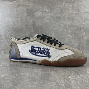 May include: A low-top sneaker featuring a white leather upper with beige suede overlays at the toe and lace area. The side displays the blue embroidered text Von Dutch. It includes black laces, a grey heel panel with a yellow logo, and a gum rubber sole.