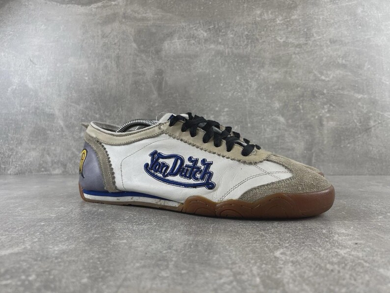 May include: A low-top sneaker featuring a white leather upper with beige suede overlays at the toe and lace area. The side displays the blue embroidered text Von Dutch. It includes black laces, a grey heel panel with a yellow logo, and a gum rubber sole.