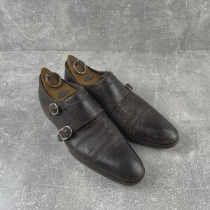 May include: A pair of dark brown leather double monk strap shoes with silver buckles. The shoes have a worn appearance and are placed on a gray surface. Wooden shoe trees are inside the shoes.