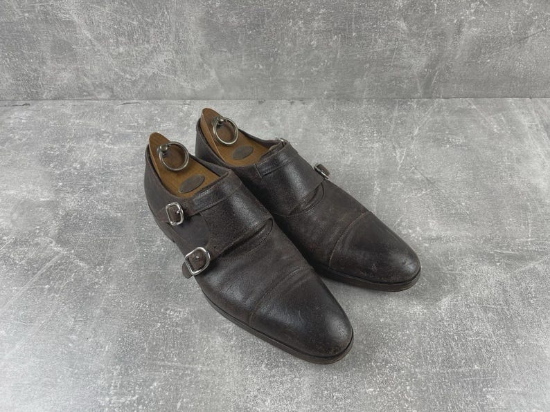 May include: A pair of dark brown leather double monk strap shoes with silver buckles. The shoes have a worn appearance and are placed on a gray surface. Wooden shoe trees are inside the shoes.