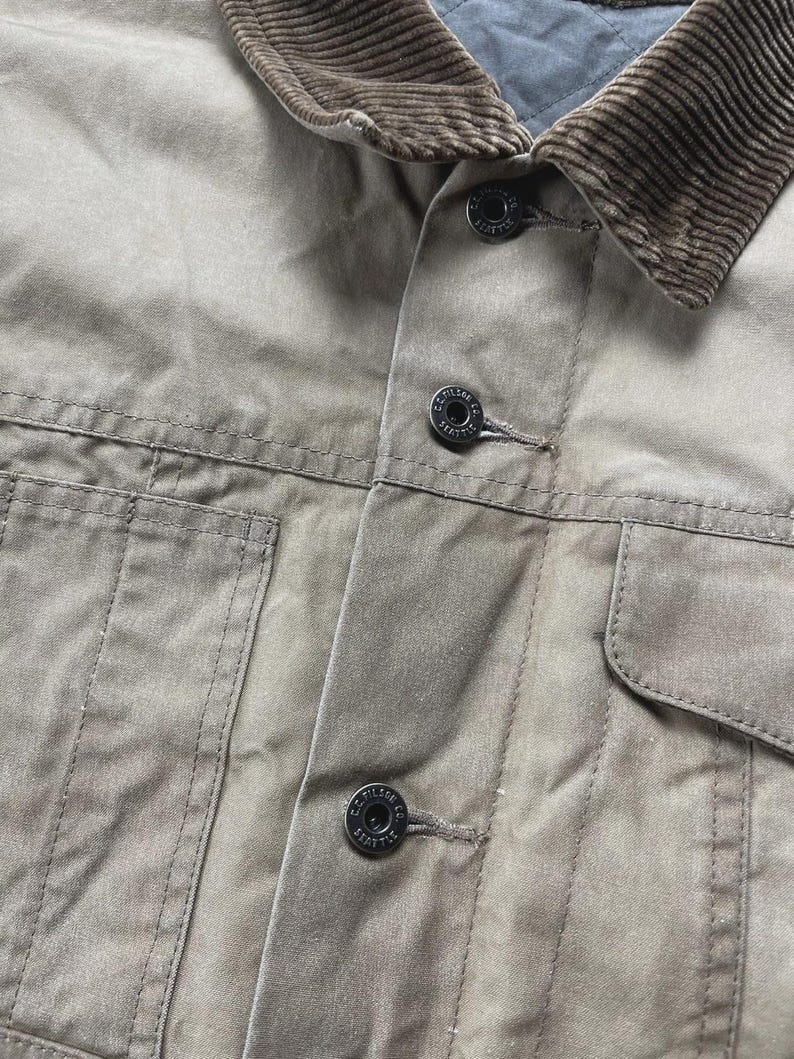 Puede incluir: Una chaqueta de color tostado con cuello de pana marr&oacute;n y forro interior acolchado. La parte delantera incluye una tapeta con botones negros grabados con el texto C.C. FILSON CO. SEATTLE, junto con dos grandes bolsillos frontales de parche.