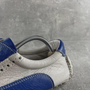 May include: Close-up of a blue and white leather trainer. The shoe has blue detailing on the heel and toe, with white leather panels. The design includes decorative perforations and white laces. A metal shoe tree is inserted.