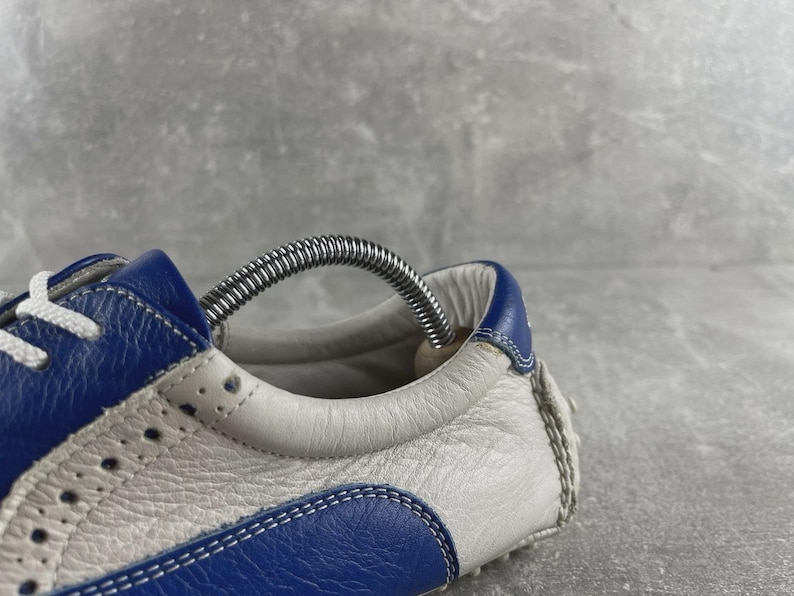 May include: Close-up of a blue and white leather trainer. The shoe has blue detailing on the heel and toe, with white leather panels. The design includes decorative perforations and white laces. A metal shoe tree is inserted.
