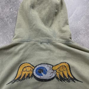 May include: A sage green hoodie featuring a distressed winged eye graphic on the back. The eye has a blue iris and yellow wings. The hoodie has a drawstring hood and a relaxed fit.