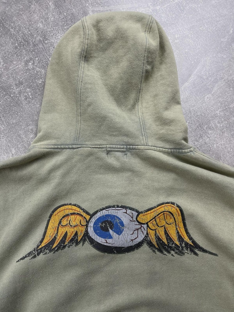 May include: A sage green hoodie featuring a distressed winged eye graphic on the back. The eye has a blue iris and yellow wings. The hoodie has a drawstring hood and a relaxed fit.