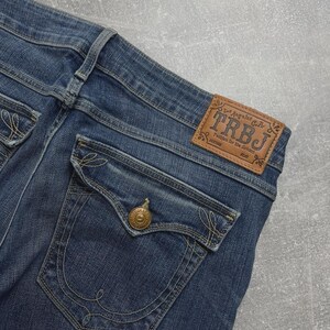 May include: Blue denim jeans featuring a tan leather patch on the waistband with the text TRBJ Los Angeles C.A. Fashion for the Senses. The back pocket has a flap with a gold-toned metal button and decorative tan stitching in a swirl pattern.