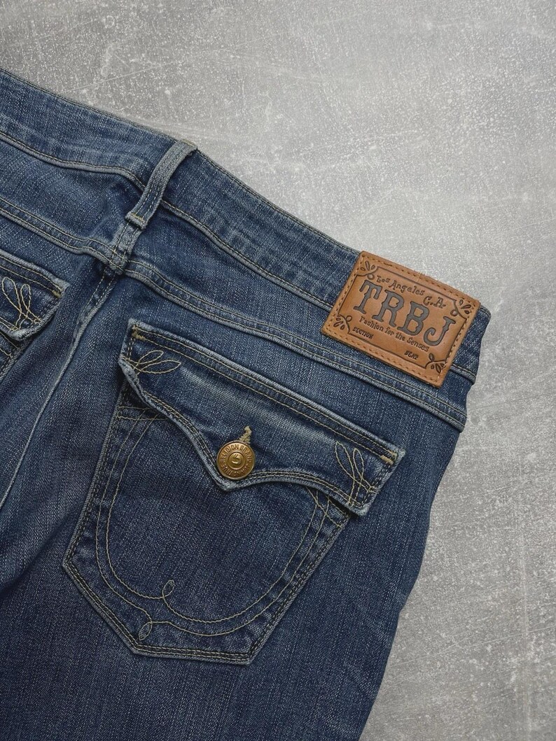 May include: Blue denim jeans featuring a tan leather patch on the waistband with the text TRBJ Los Angeles C.A. Fashion for the Senses. The back pocket has a flap with a gold-toned metal button and decorative tan stitching in a swirl pattern.